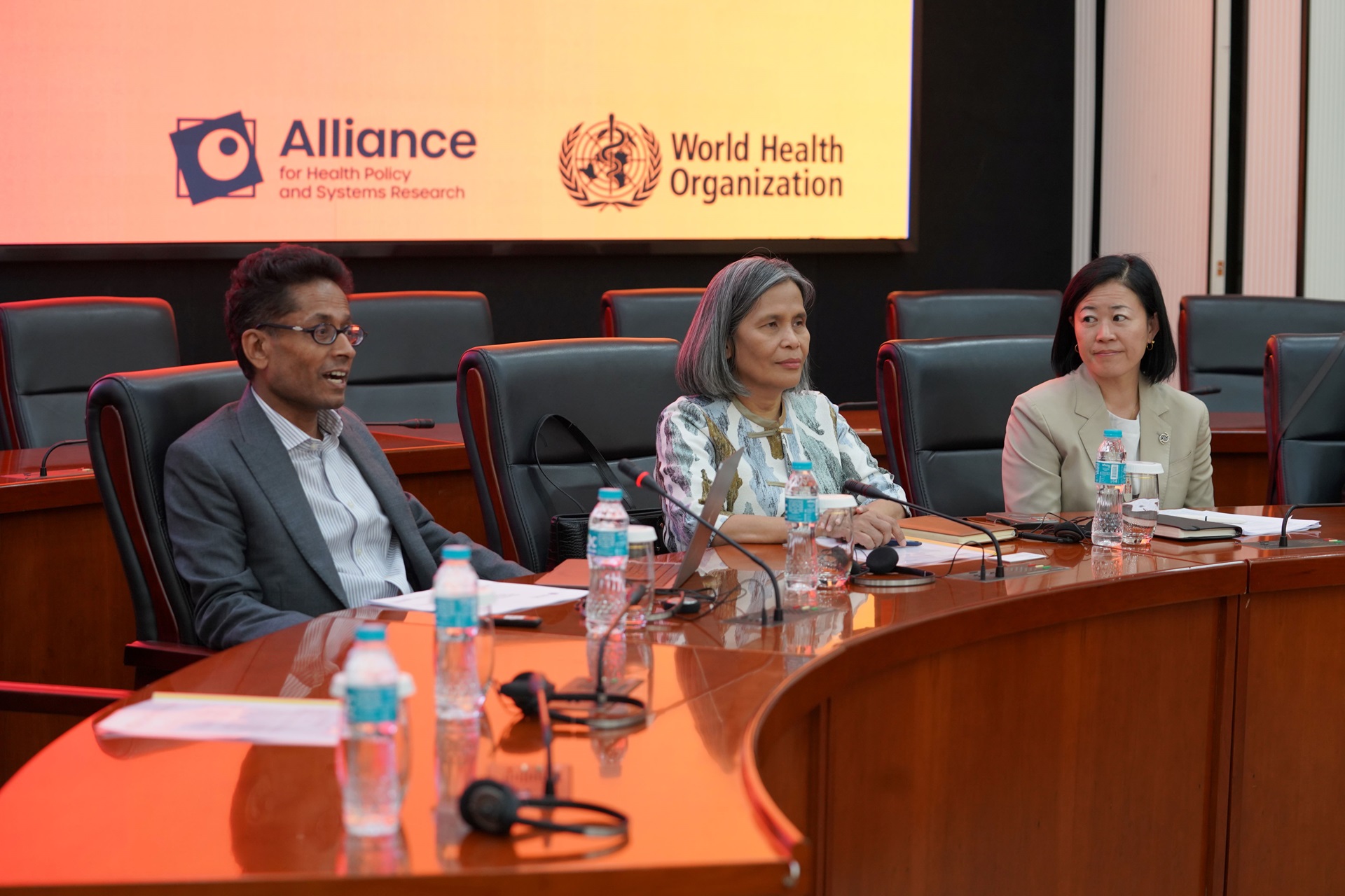 Participants seated at a conference table during a WHO meeting. Screen shows “Alliance for Health Policy and Systems Research”.