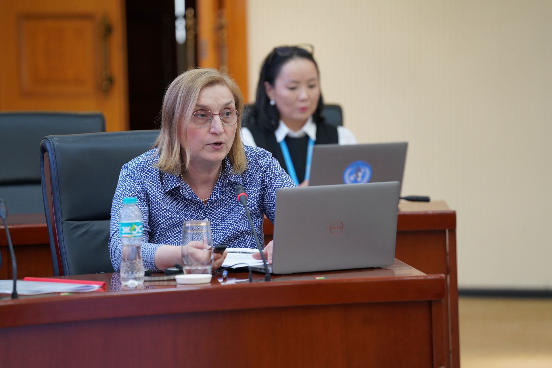 Participant speaks on a microphone while using a laptop at a meeting table with documents. Another attendee in the background.