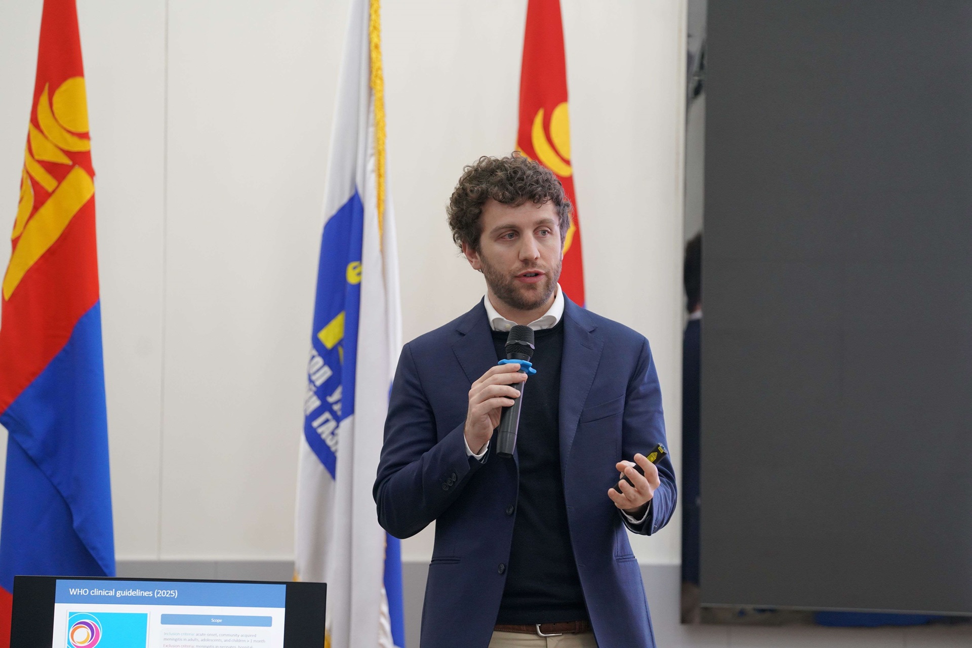 A man speaks while holding a mic. Mongolia flags are behind him.