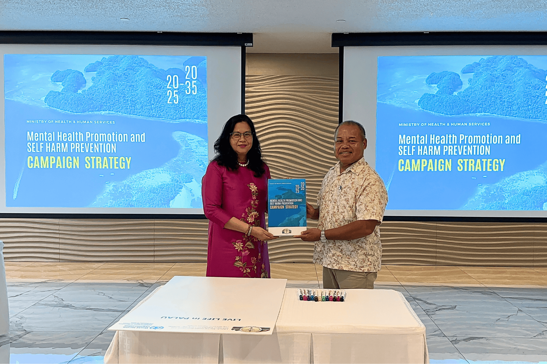A man and a woman hold a publication. Behind is text that says mental health promotion and self-harm prevention campaign strategy