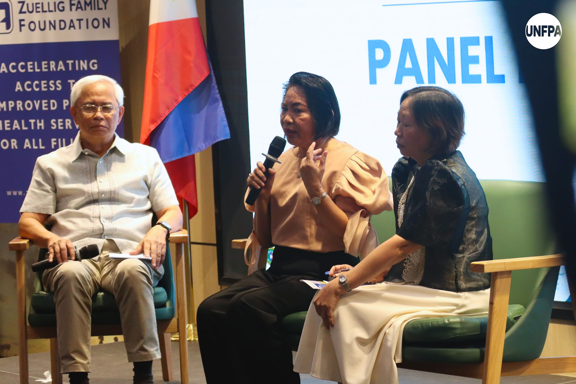 Three speakers seated on stage during a panel discussion with microphones and notes, beside a Philippine flag.