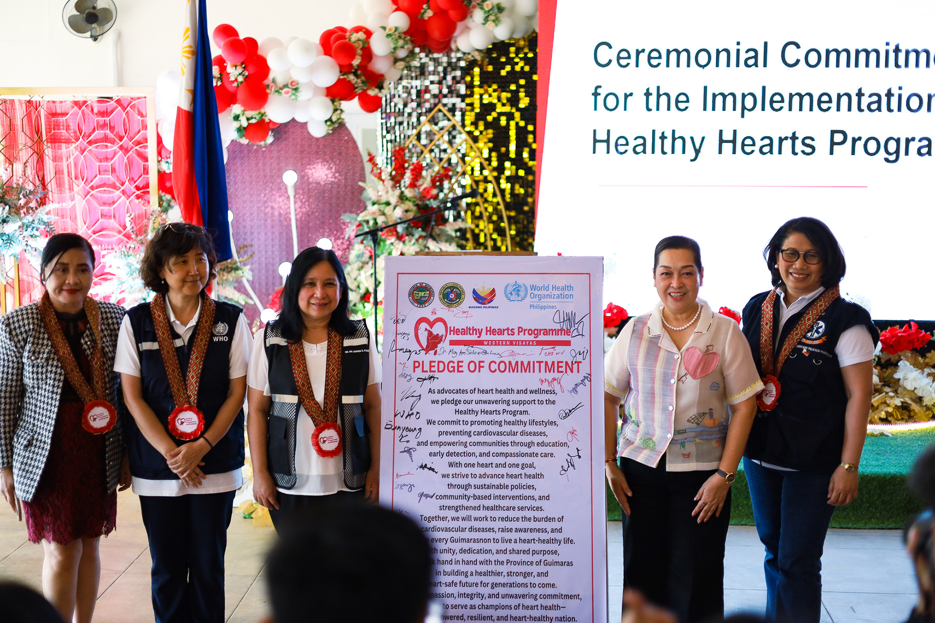 Five individuals stand before a 'Pledge of Commitment' poster at a Healthy Hearts Program implementation event, with festive decorations and event banner in the background.