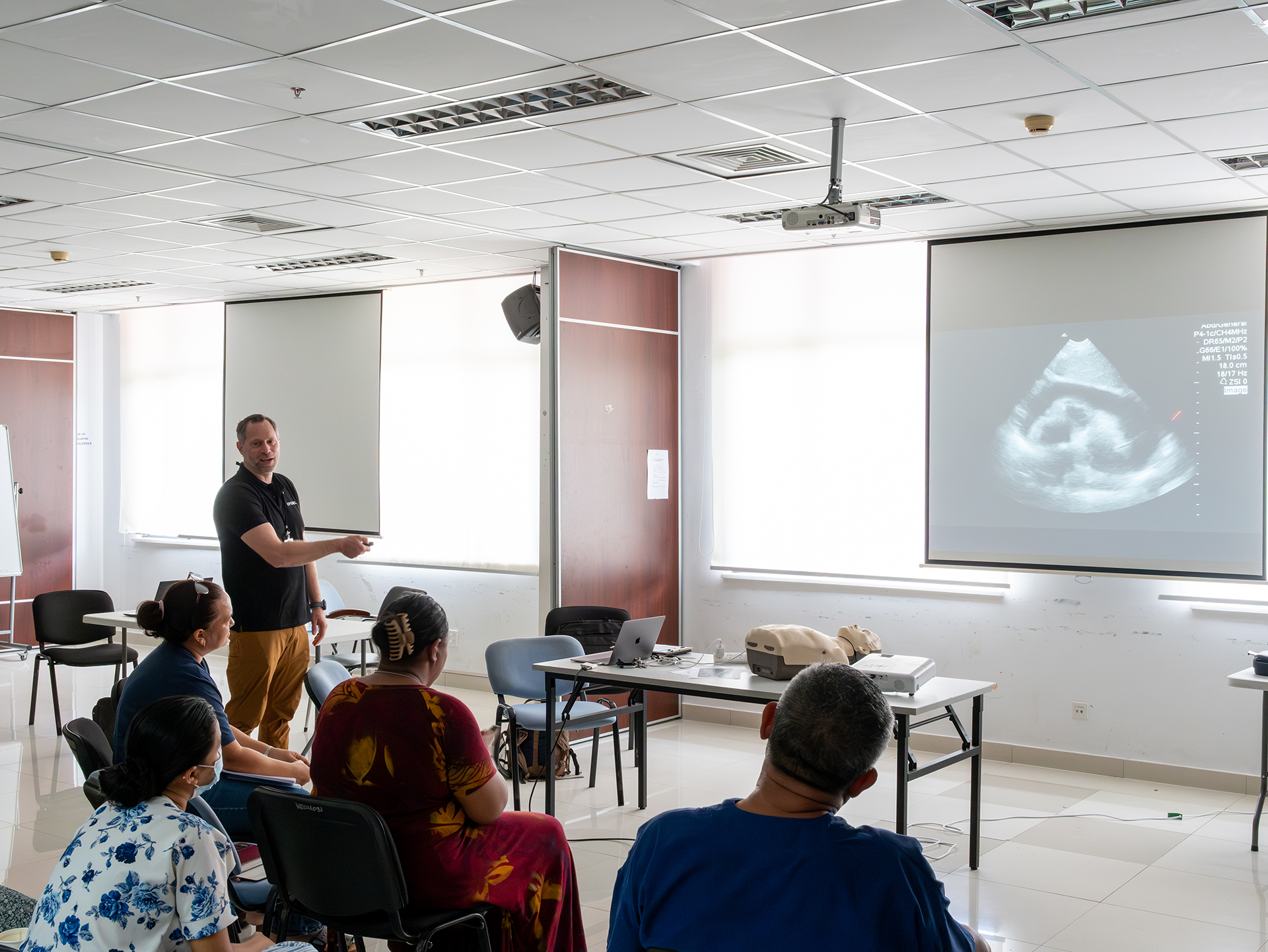 A doctor is standing and gesturing toward a projected ultrasound image on a screen, while participants sit in a classroom setting with equipment on a table.