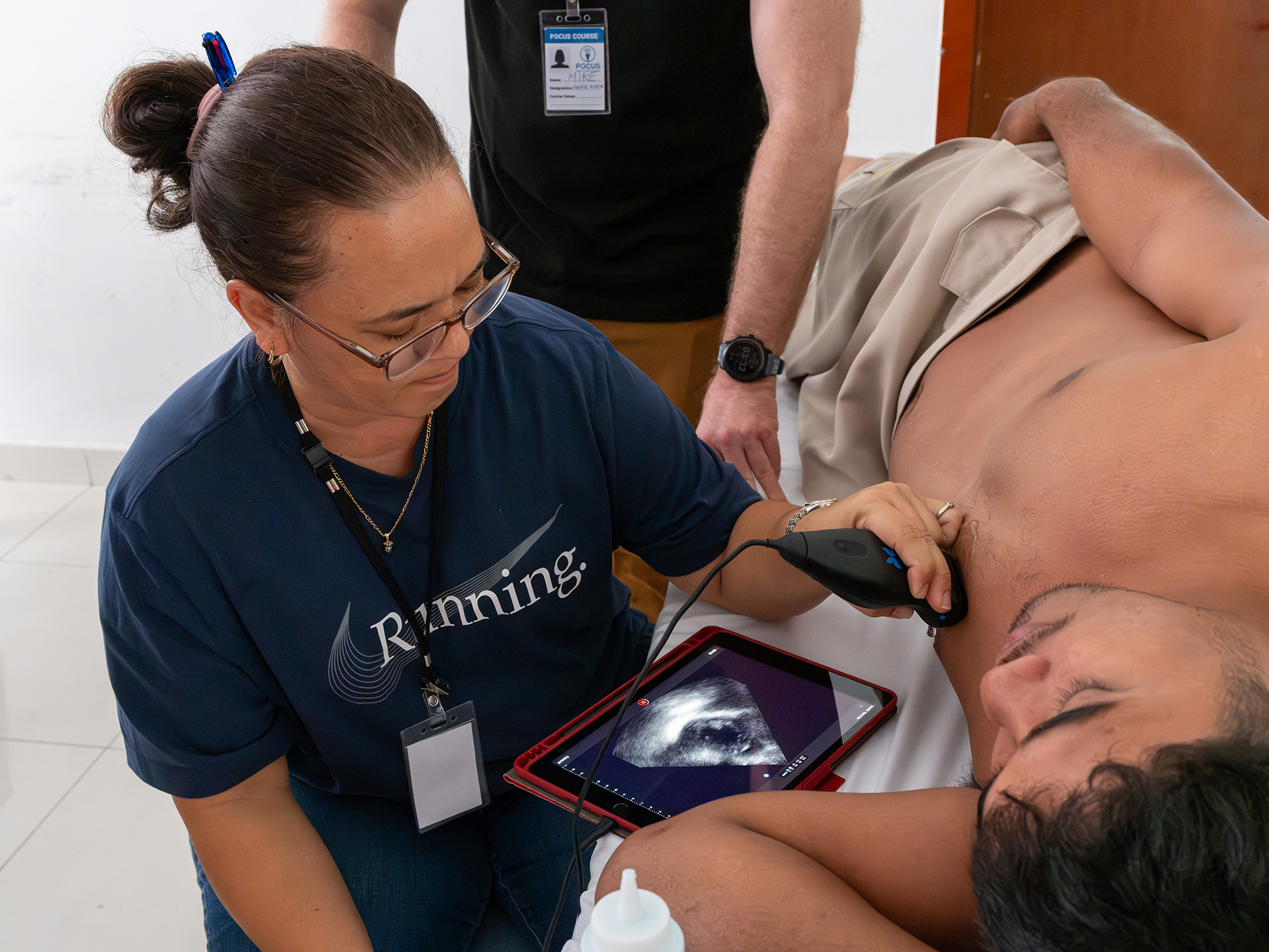 A doctor in a blue shirt practices using a portable ultrasound device on a male volunteer’s chest. The ultrasound image is visible on a tablet screen.