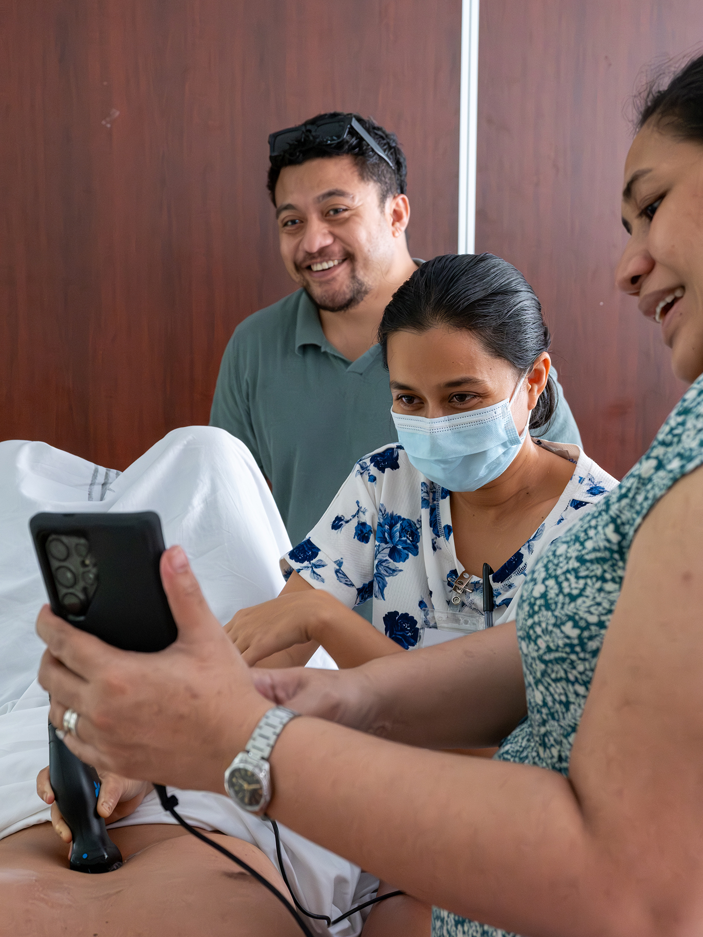 Doctors conduct an ultrasound scan on a male volunteer's abdomen. One doctor holds the probe while the two others observe closely. The volunteer lies down with a white sheet covering the lower body.