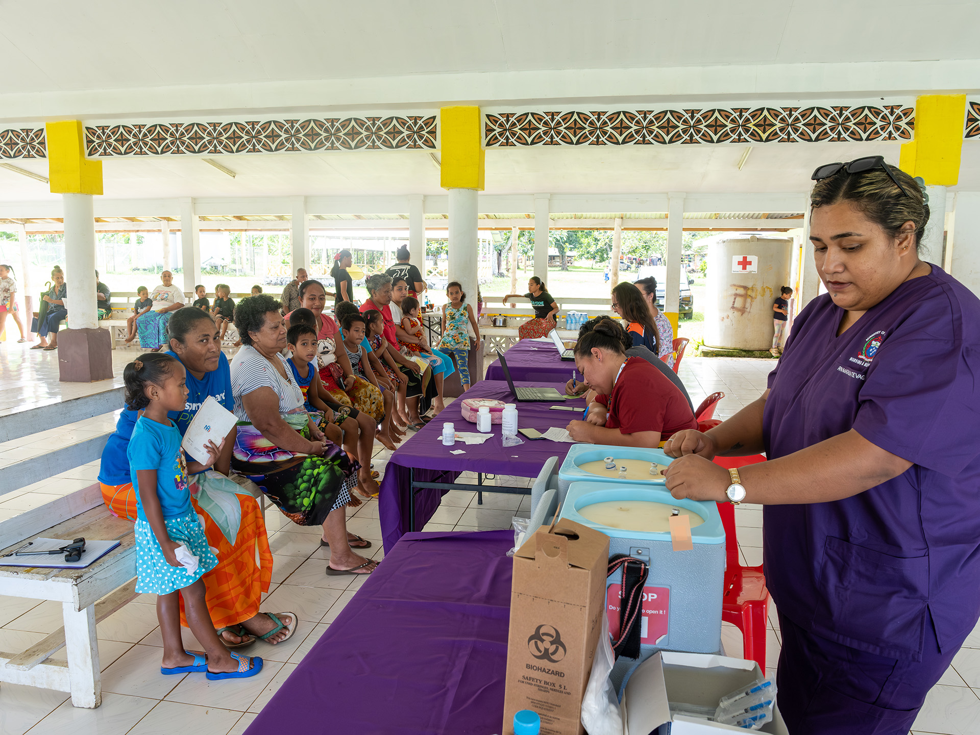 Health workers prepare medical supplies while community members wait during an outreach session in a village hall.