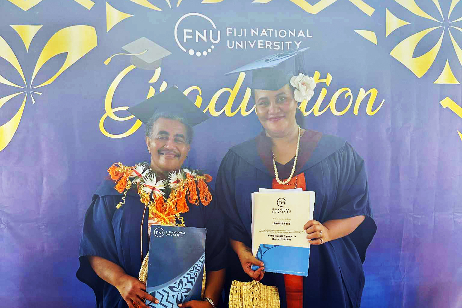 Two graduates in academic gowns and caps standing in front of a Fiji National University backdrop that reads ‘Graduation,’ holding their certificates.