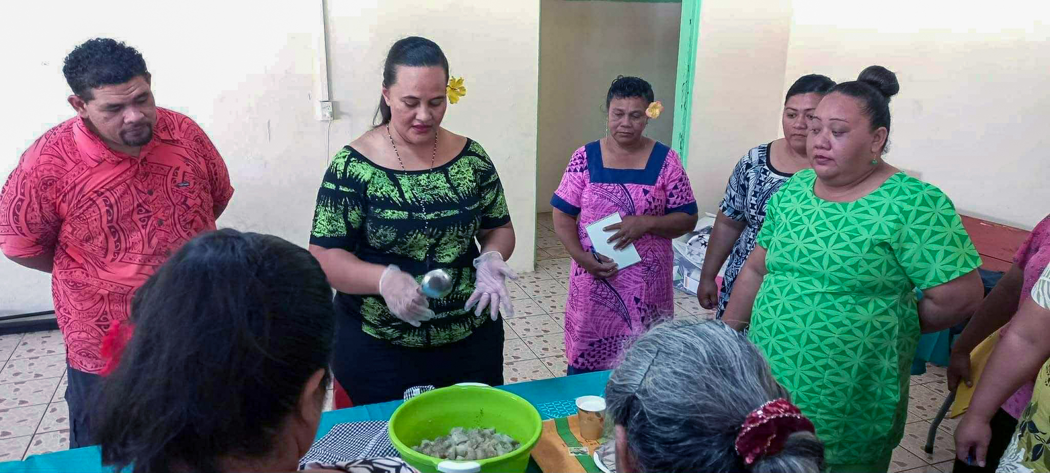 Community outreach session on healthy eating and nutrition, with a facilitator demonstrating food preparation using fresh ingredients to a group gathered around a table.
