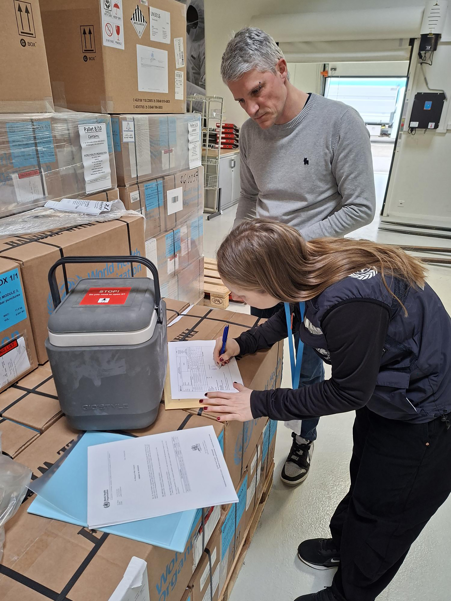 Two staff review and sign shipment documents beside stacked WHO-labeled boxes and a transport cooler in a warehouse.