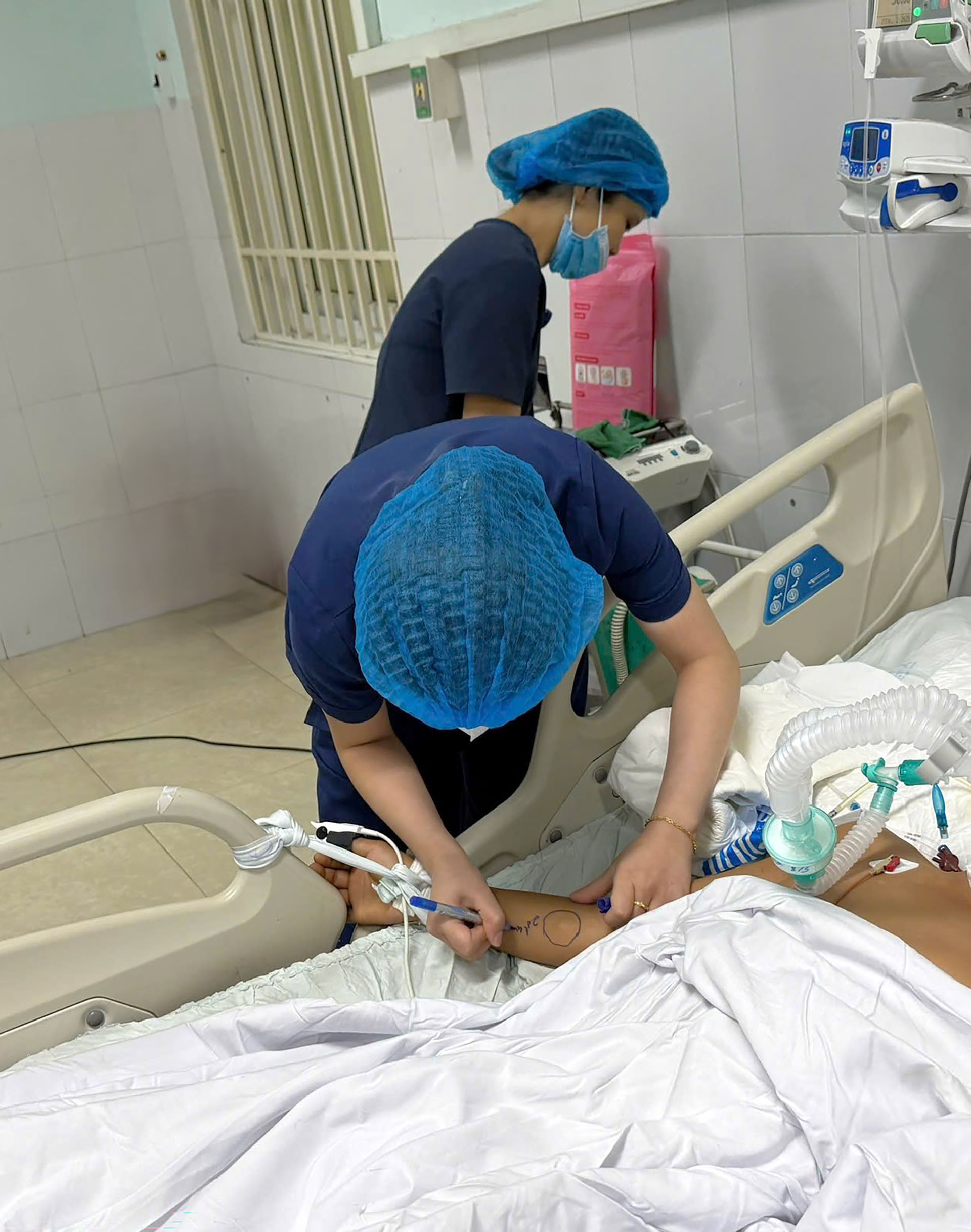 Healthcare workers adjust medical tubing and mark a patient’s arm beside a hospital bed with monitoring equipment.