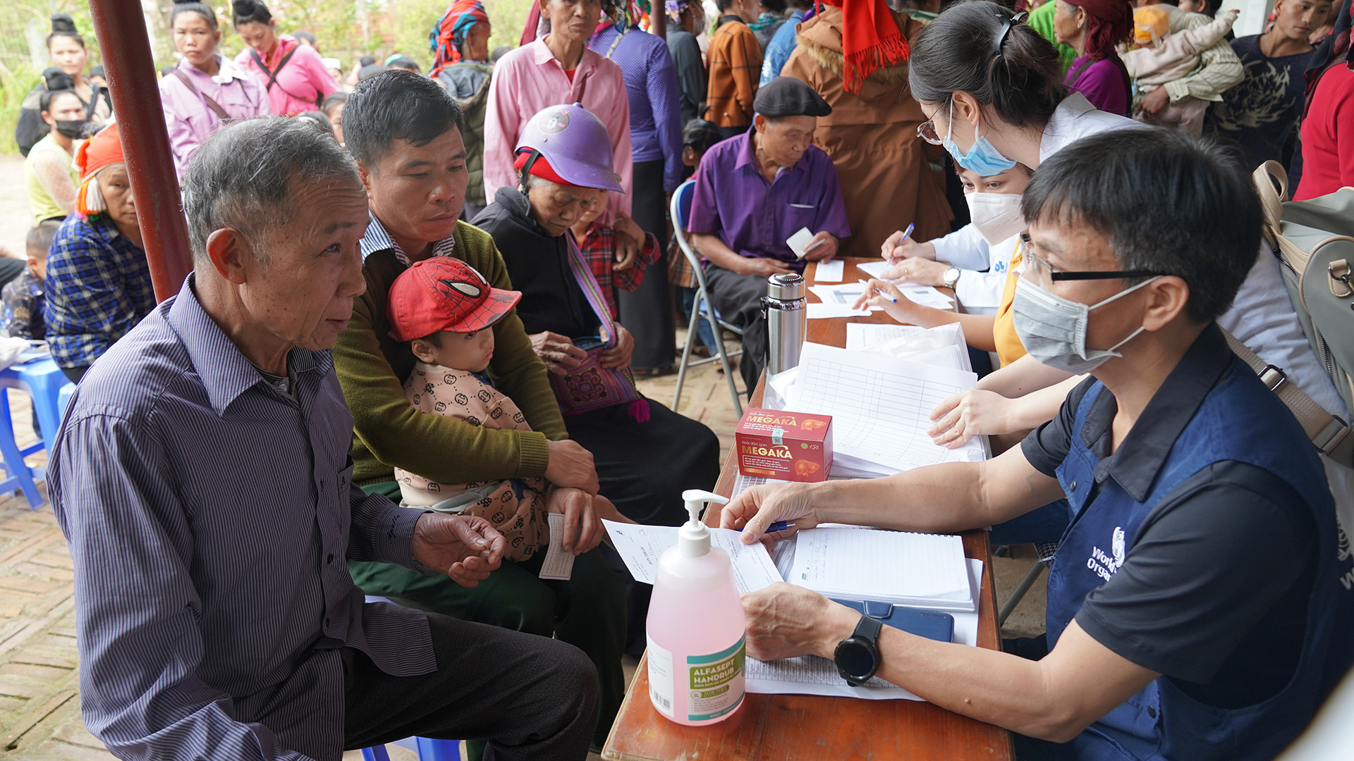 People gathered at a registration table. Individuals at a table are filling out forms. A person holds a child while others wait in line in the background.