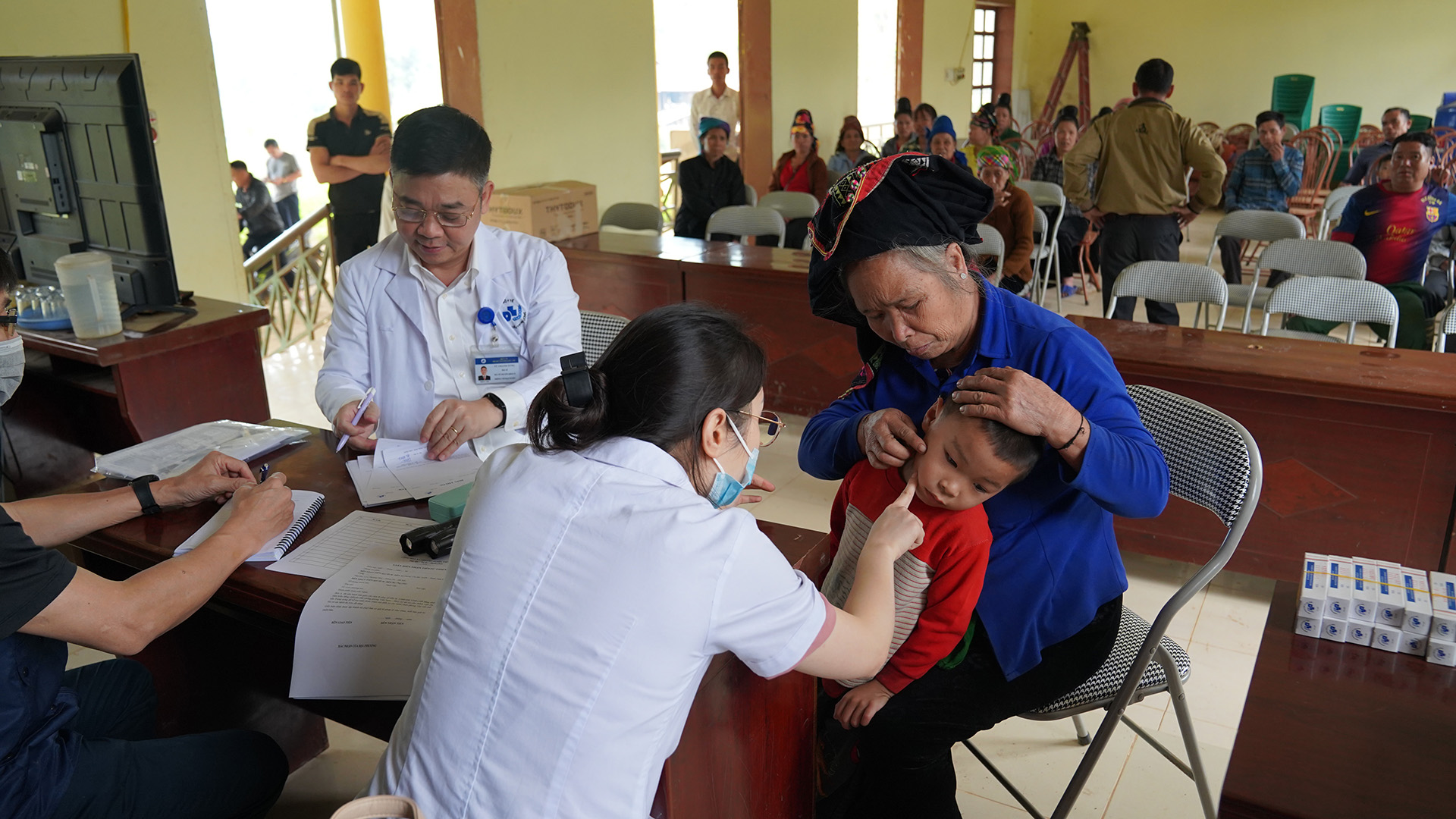A masked medical professional in a white examines a young child wearing a red and beige sweater, held by an adult in blue attire with a headscarf. Another healthcare worker writes notes at a table, while several people wait seated in the background.