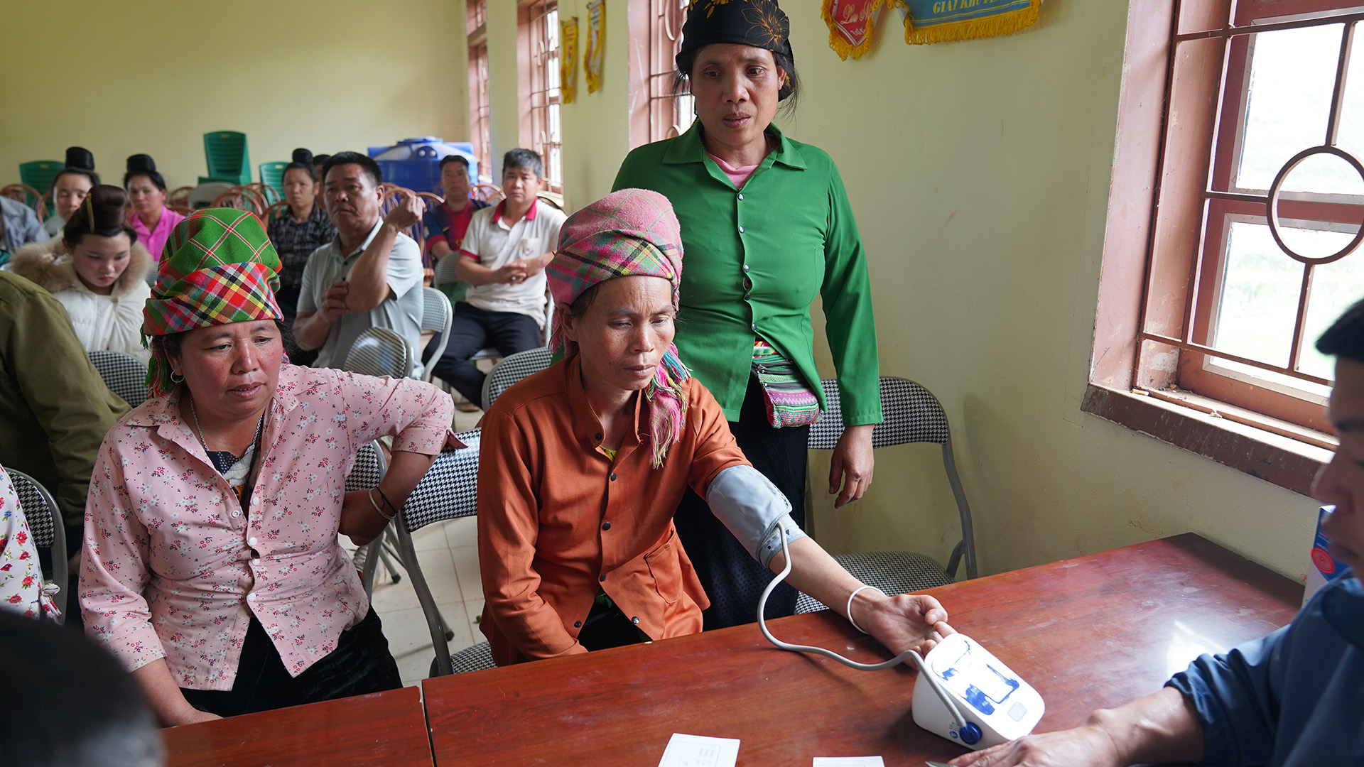 A person in an orange shirt and colourful headscarf has their blood pressure checked using an electronic device. A person in a green shirt stands nearby, while others sit in rows of chairs in the background.