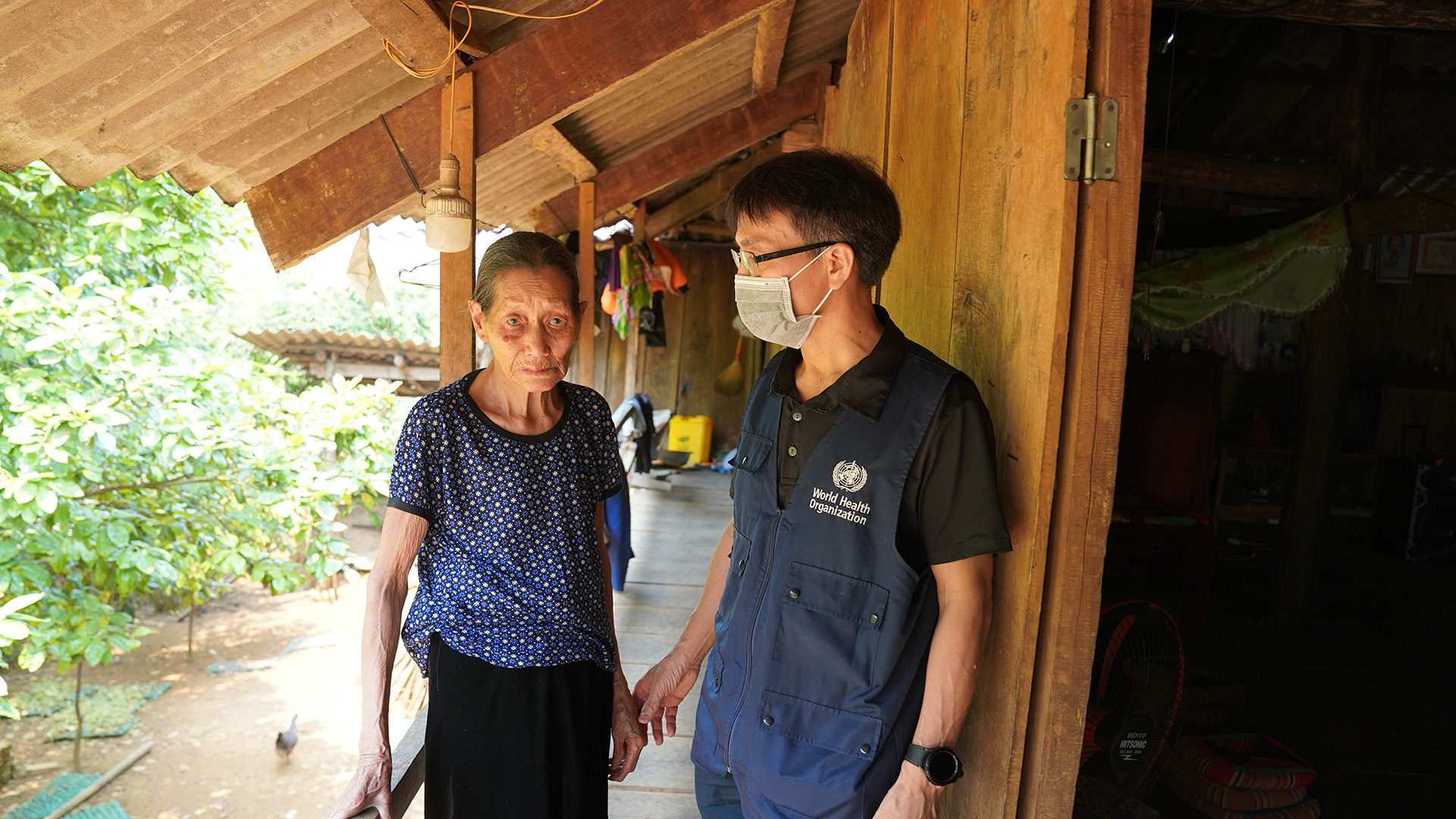 An elderly person in a blue patterned shirt and black trousers stands on the porch of a wooden house, speaking with a World Health Organization worker wearing a dark shirt and WHO vest.