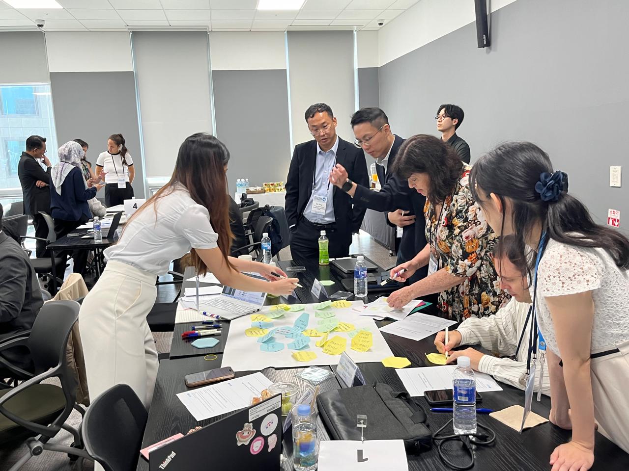 People dressed in formal clothes work together and place sticky notes with writing on a large piece of paper. Water bottles, open laptops, notes and mobile phones are scattered all over the table, indicating a productive meeting.