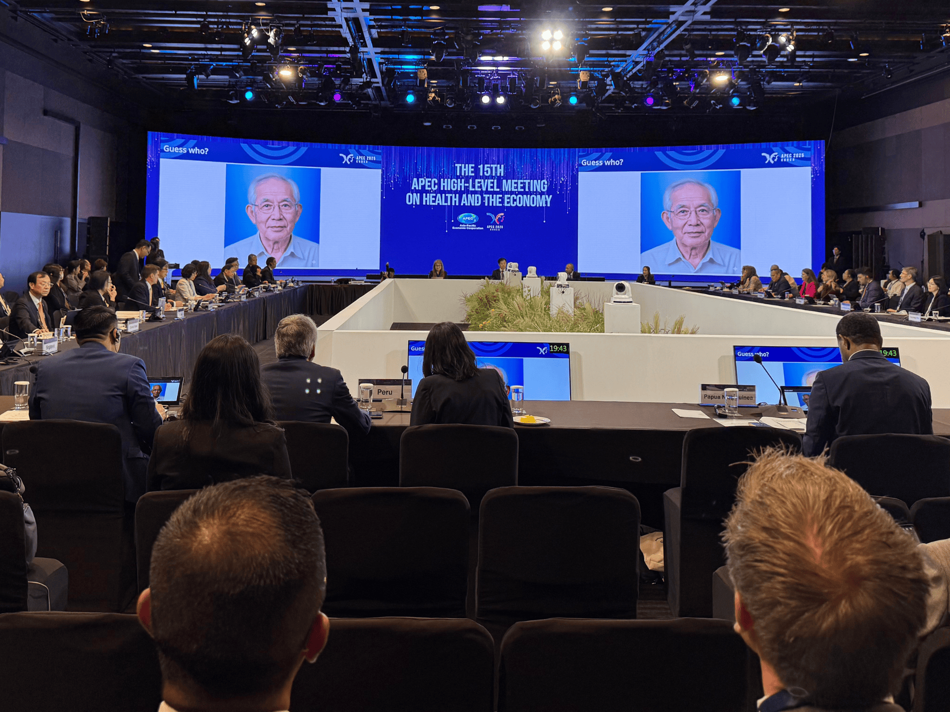 A stage in the background with a photo of an older person on screen, audience looking in the foreground