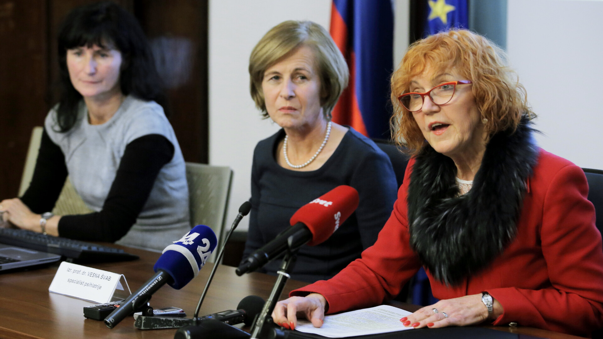 A smartly dressed woman in red jacket and glasses speaks to an audience out of frame. Two other woman is sitting at the same table listening.