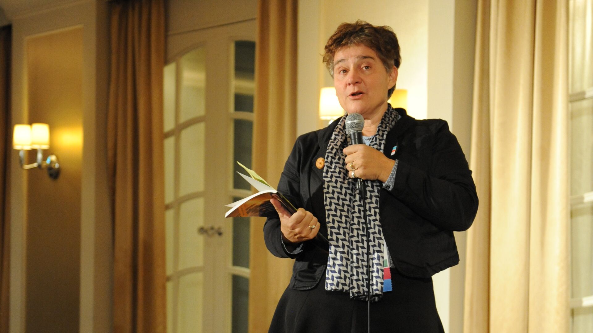 A woman wearing a scarf around her neck and holding a booklet in one hand and a microphone in the other, is speaking to an audience out of frame.