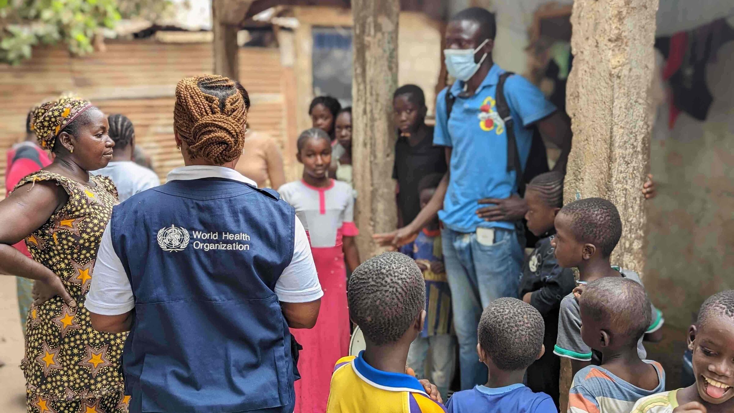 A WHO officer seen from behind, talks to a group of adults and children in a village setting.