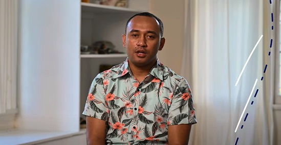 Video snapshot of a man sitting on a chair talking.