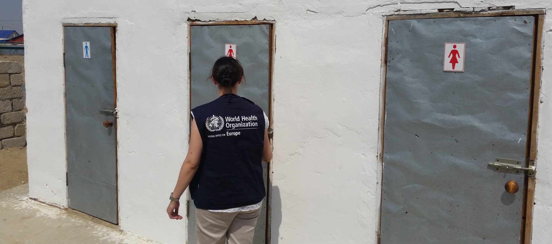 A person wearing a World Health Organization Europe vest approaches a public toilet door.