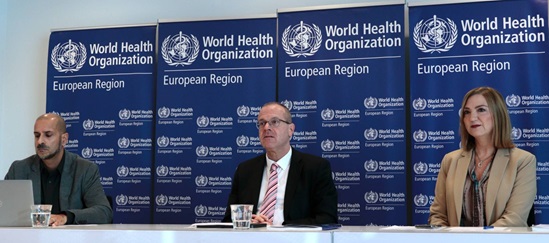 WHO representatives sit at a table in front of WHO banner during a press briefing