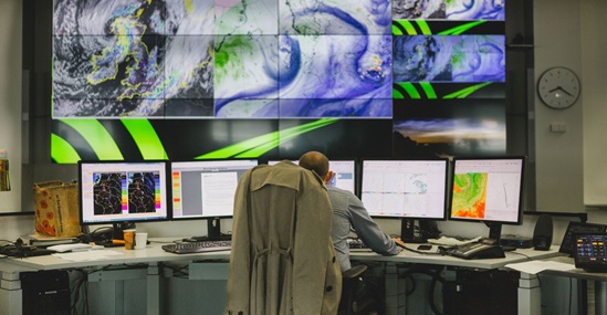 A person monitors complex climate and weather patterns in a high-tech control room equipped with large and small digital screens showing maps and forecasts