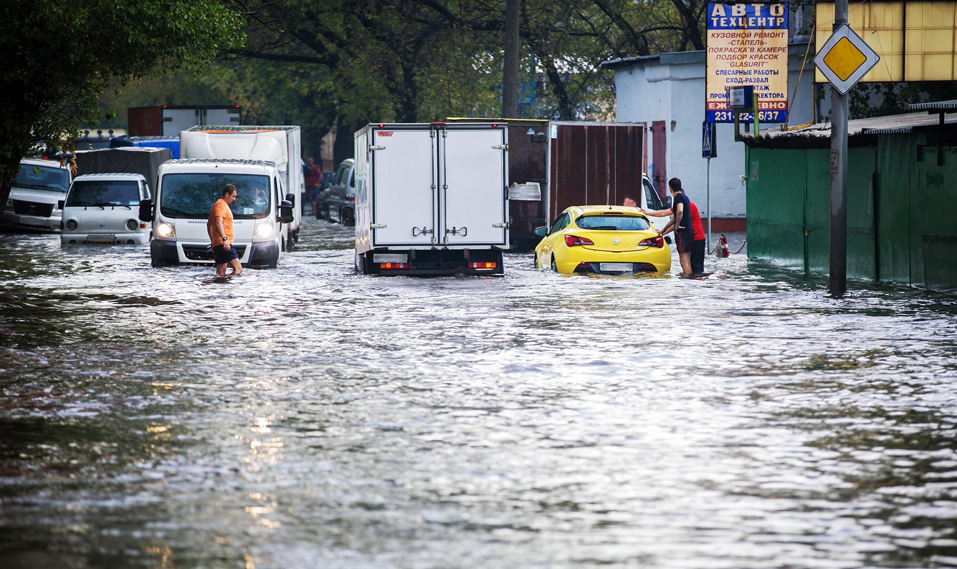 A road is flooded; cars and people move and wade through the water, trying to deal with the situation.
