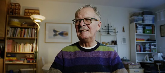 An older man in glasses and a striped sweater smiles in a cozy room