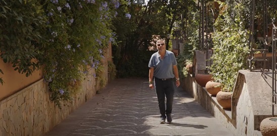 A man walks alone down a sunlit stone pathway bordered by lush greenery and terracotta planters