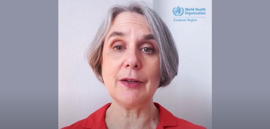 A woman with short gray hair in a red blouse speaks on camera against a plain wall