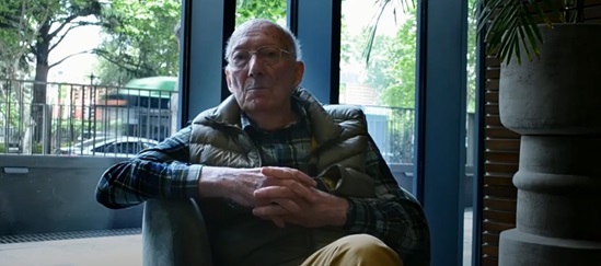 An older man in glasses and a vest sits quietly indoors near a large window