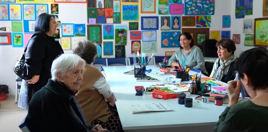 A group of older adults sits around a table filled with art supplies in a brightly decorated room, engaging in an arts and crafts session.