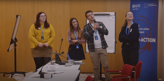 Meeting participants standing, one of them holding a mike, all are looking in the same direction, possibly a screen