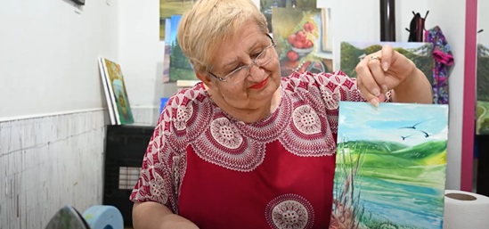 Smiling elderly woman shows a hand-painted picture of a bright landscape with hills, water, and birds